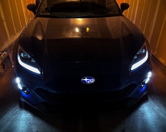 May include: A blue Subaru BRZ with white headlights and fog lights. The car is parked in a garage with a yellow wall in the background.