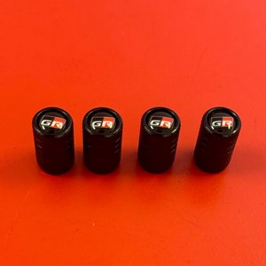 Puede incluir: Cuatro tapones de v&aacute;lvula de neum&aacute;tico negros con un logotipo "GR" rojo y blanco.