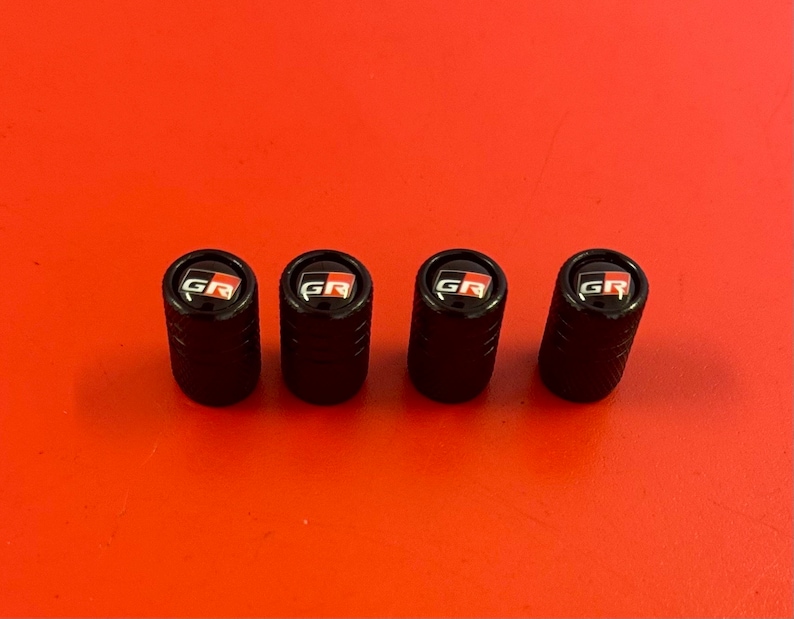 Puede incluir: Cuatro tapones de v&aacute;lvula de neum&aacute;tico negros con un logotipo "GR" rojo y blanco.