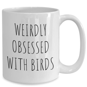May include: A white ceramic mug featuring the black printed text WEIRDLY OBSESSED WITH BIRDS in a simple sans-serif font. The mug has a smooth finish, a cylindrical body, and a curved handle on the right side, providing a clean and functional design.