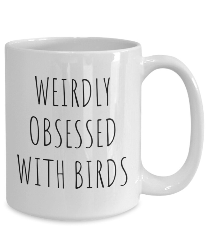 May include: A white ceramic mug featuring the black printed text WEIRDLY OBSESSED WITH BIRDS in a simple sans-serif font. The mug has a smooth finish, a cylindrical body, and a curved handle on the right side, providing a clean and functional design.