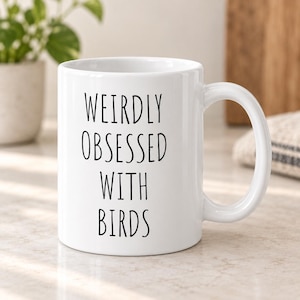 May include: A white ceramic mug featuring the black printed text WEIRDLY OBSESSED WITH BIRDS in a simple sans-serif font. The mug has a standard cylindrical shape with a curved handle on the right side and a smooth, glossy finish throughout the exterior.