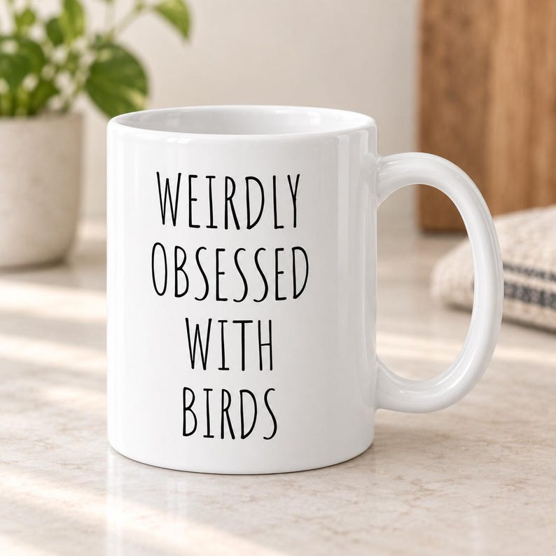 May include: A white ceramic mug featuring the black printed text WEIRDLY OBSESSED WITH BIRDS in a simple sans-serif font. The mug has a standard cylindrical shape with a curved handle on the right side and a smooth, glossy finish throughout the exterior.