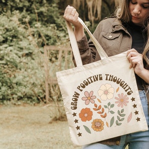 May include: A beige tote bag with the text "Grow Positive Thoughts" in brown lettering. The bag features a floral design with pink, orange, and green flowers.