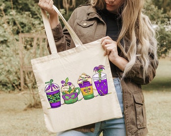 May include: A white canvas tote bag with a colorful design of four different drinks. The drinks are decorated with Mardi Gras beads, a mask, and a palm tree.