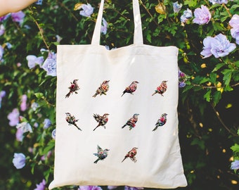 May include: Cream-colored tote bag featuring colorful floral bird designs. The birds are depicted in various poses, with detailed floral patterns on their wings and bodies. The bag has long handles and is held up against a backdrop of green foliage and purple flowers.