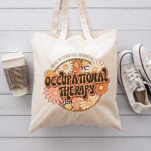 May include: Cream-colored tote bag with the words "OCCUPATIONAL THERAPY" in a retro font, surrounded by daisies and smiley faces. A white coffee cup with a knitted sleeve and white sneakers are also in the image.