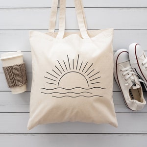 May include: A white canvas tote bag with a black line drawing of a sun setting over the ocean. The bag has two handles and is sitting on a white wooden surface. There is a white coffee cup with a brown knitted sleeve and two white trainers on the surface.
