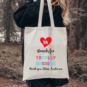 May include: A white canvas tote bag with a red heart that says "TA" inside. The text below the heart says "stands for TOTALLY AWESOME thank you Miss Anderson".