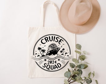 May include: Cream-colored tote bag with black graphic of a cruise ship, palm trees, and text that reads "CRUISE 2024 SQUAD". A beige hat rests above the bag. The bag has two handles and is made of a canvas-like material.