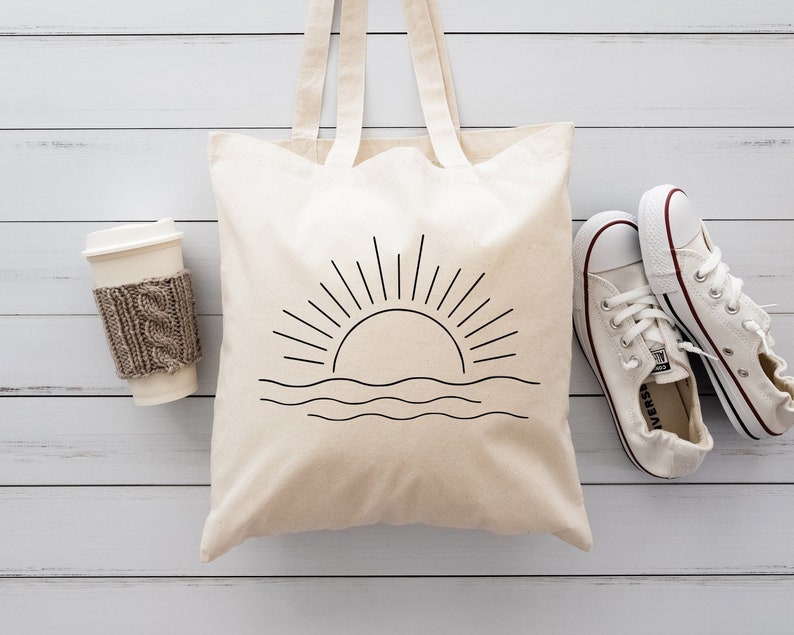 May include: A white canvas tote bag with a black line drawing of a sun setting over the ocean. The bag has two handles and is sitting on a white wooden surface. There is a white coffee cup with a brown knitted sleeve and two white trainers on the surface.