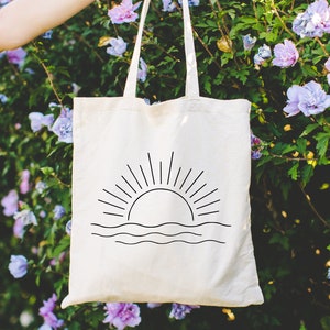 May include: A white canvas tote bag featuring a black line drawing of a sun rising over the ocean waves.