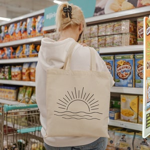 May include: A woman wearing a white hoodie is shopping in a supermarket. She is carrying a beige tote bag with a black sun and waves design. The bag is hanging from her shoulder. The supermarket shelves are stocked with various food items, including cereal boxes and bags of crisps.