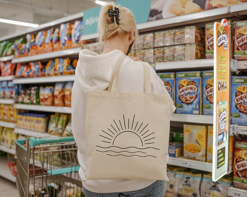 May include: A woman wearing a white hoodie is shopping in a supermarket. She is carrying a beige tote bag with a black sun and waves design. The bag is hanging from her shoulder. The supermarket shelves are stocked with various food items, including cereal boxes and bags of crisps.