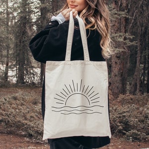 May include: A white canvas tote bag with black straps and a simple line drawing of a sunset with waves. The bag is being held by a person in a woodland setting.
