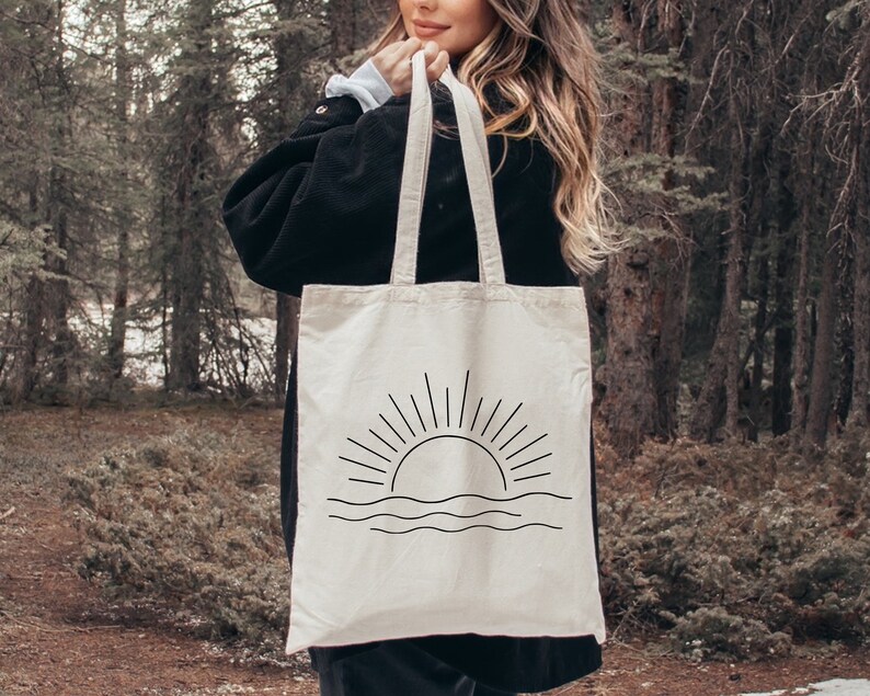 May include: A white canvas tote bag with black straps and a simple line drawing of a sunset with waves. The bag is being held by a person in a woodland setting.