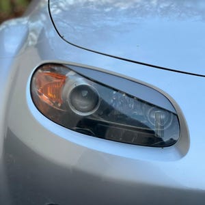 May include: Close-up of a silver car's front, highlighting the headlight. The headlight has an orange indicator and a clear lens. The car's body has a smooth, metallic finish.