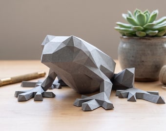 May include: A gray, low-poly geometric frog figurine with sharp, angular facets. The sculpture features a stylized, multi-faceted surface design, showcasing a modern, segmented appearance that defines its head, body, and splayed limbs.