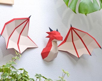 May include: A red and white paper dragon head and wings are mounted on a white wall. The dragon's head is in the center, with a wing on each side. The wings and head have black accents.