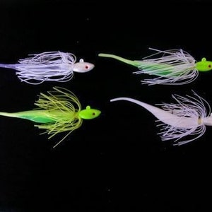 May include: Four fishing lures with a streamlined design are displayed against a black background. Each lure features a brightly colored head, a long, thin tail, and a skirt of fine, flowing fibers. The color palette includes white, green, yellow, and purple.