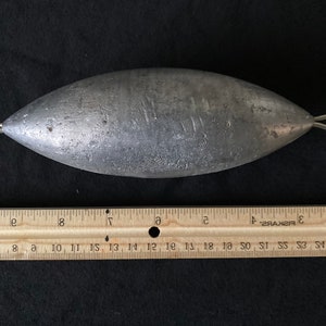May include: A silver-colored, torpedo-shaped fishing weight with two loops at the ends. The weight is 9 inches long and is measured against a wooden ruler.
