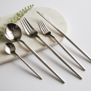 May include: A set of five silver-toned flatware pieces, including a knife, two forks, and two spoons, arranged on a white marble surface.