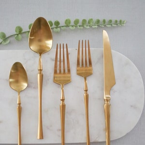 May include: A set of five gold-toned flatware pieces, including a spoon, two forks, and a knife, arranged on a white marble surface.