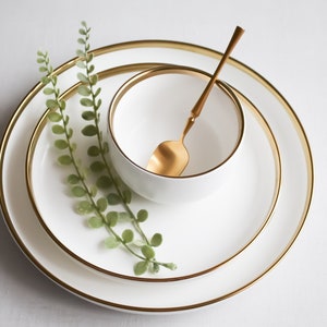 May include: A set of three white ceramic plates with gold trim. The plates are stacked on top of each other, with a small white bowl and a gold spoon in the center. A sprig of green foliage is placed on the plates.