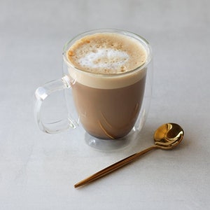 May include: A glass mug with a double wall design filled with a light brown coffee drink with a white foam top. A gold-colored metal spoon is next to the mug.