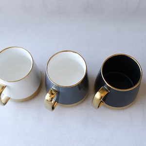 May include: Three ceramic mugs with gold rims and handles. One mug is white, one is gray, and one is black.