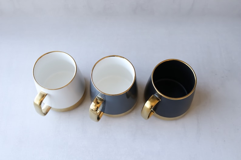 May include: Three ceramic mugs with gold rims and handles. One mug is white, one is gray, and one is black.