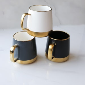 May include: Three ceramic mugs with gold rims. One mug is white, and two mugs are dark gray.