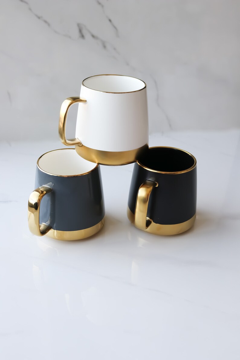 May include: Three ceramic mugs with gold rims. One mug is white, and two mugs are dark gray.