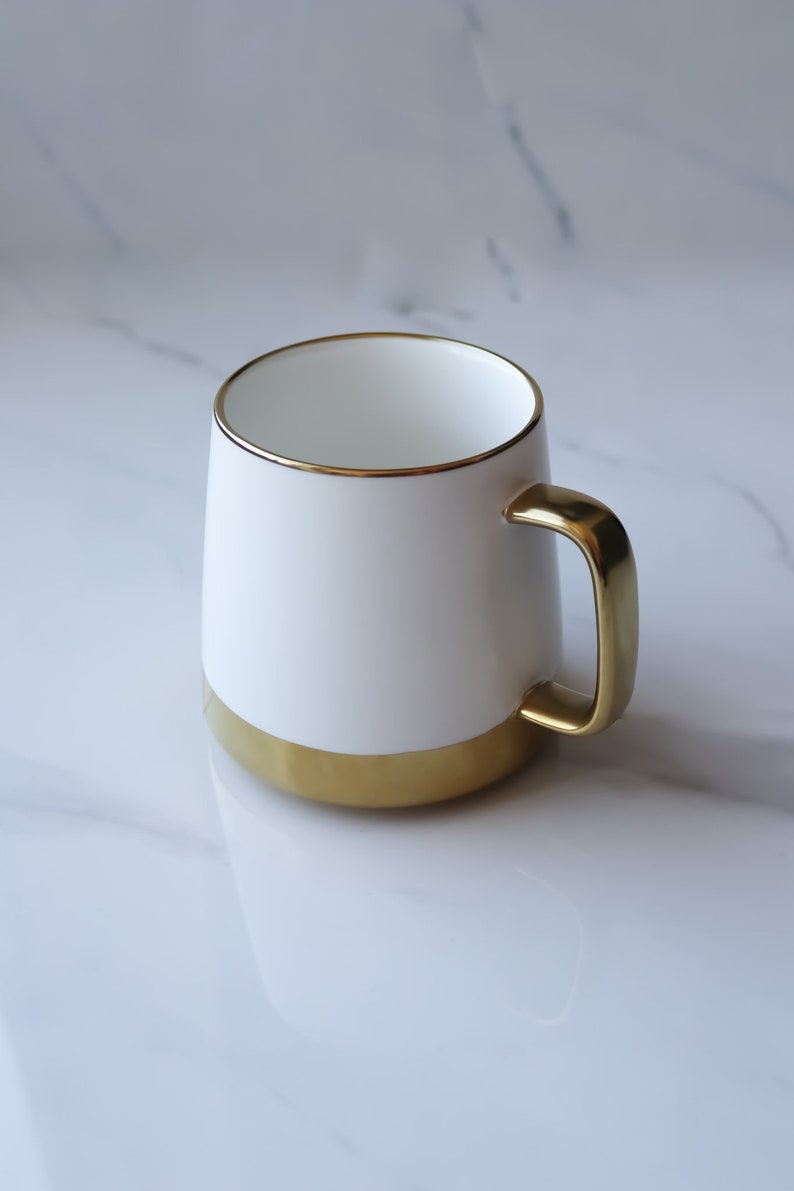 May include: A white ceramic mug with a gold rim and base. The mug has a gold handle.