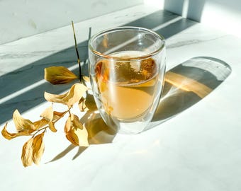May include: A clear glass double-walled mug filled with a light amber liquid, sitting on a white marble surface with dried leaves scattered around it.