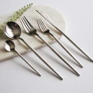 May include: A set of five silver-toned flatware pieces, including a knife, two forks, and two spoons, arranged on a white marble surface.