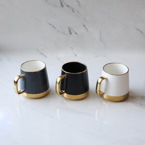 May include: Three ceramic mugs with gold rims. The mugs are black, white, and gray.