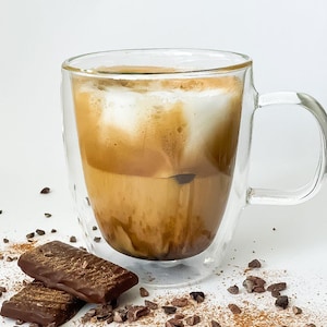 May include: A double-walled glass mug filled with a layered coffee drink, topped with a thick layer of foam. The mug is sitting on a white surface with scattered cocoa nibs and two pieces of dark chocolate.