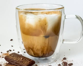 May include: A double-walled glass mug filled with a layered coffee drink, topped with a thick layer of foam. The mug is sitting on a white surface with scattered cocoa nibs and two pieces of dark chocolate.