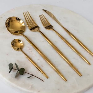 May include: A set of gold-colored flatware, including a spoon, a small spoon, two forks, and a knife, arranged on a white marble surface. The utensils have long, slender handles and are displayed with a sprig of greenery.