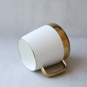 May include: White ceramic mug with a gold rim and handle. The mug is on a gray background.