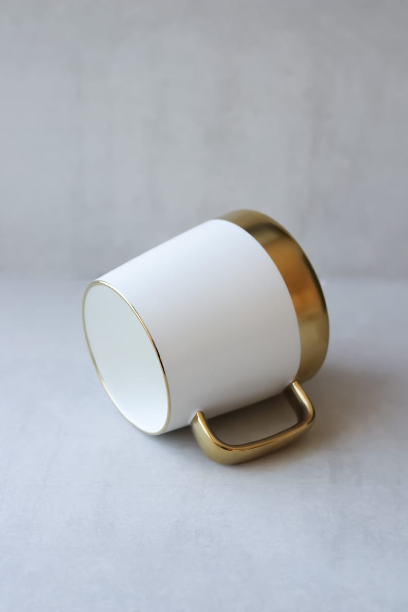 May include: White ceramic mug with a gold rim and handle. The mug is on a gray background.