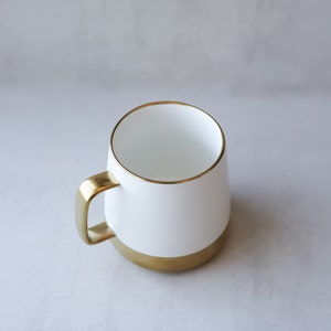May include: A white ceramic mug with a gold rim and base. The mug is empty and sitting on a gray surface.