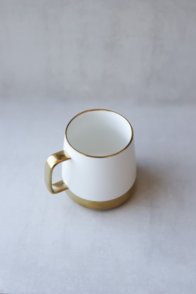 May include: A white ceramic mug with a gold rim and base. The mug is empty and sitting on a gray surface.