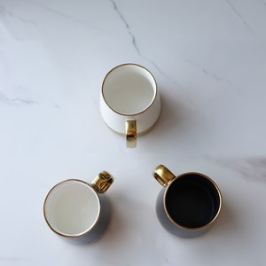 May include: Three ceramic mugs with gold trim. One mug is white, one is black, and one is gray.