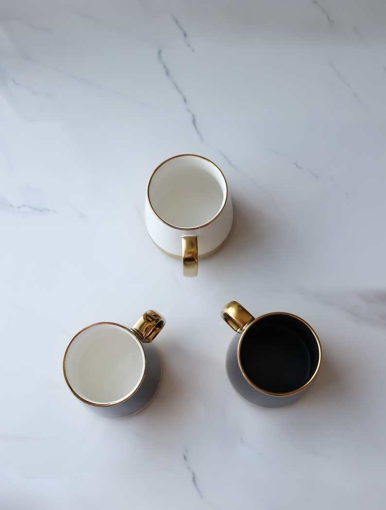 May include: Three ceramic mugs with gold trim. One mug is white, one is black, and one is gray.