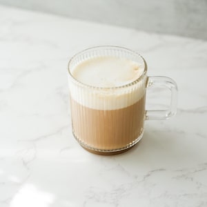 May include: A clear glass mug filled with a layered beverage. The bottom layer is a light brown color, topped with a thick layer of white foam. The mug has a handle and vertical ribbed detailing.