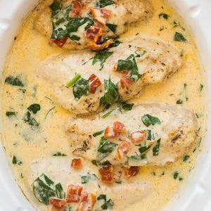 May include: Four chicken breasts cooked in a creamy yellow sauce with green spinach leaves and chopped sun-dried tomatoes inside a white oval slow cooker. The text "the RECIPE REBEL" is visible in the bottom left corner of the image.