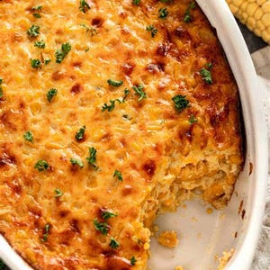 May include: A golden-brown baked corn casserole in a white oval ceramic baking dish. The dish features a textured, browned top surface garnished with small green parsley flakes, with a portion removed to reveal the dense, yellow corn interior.