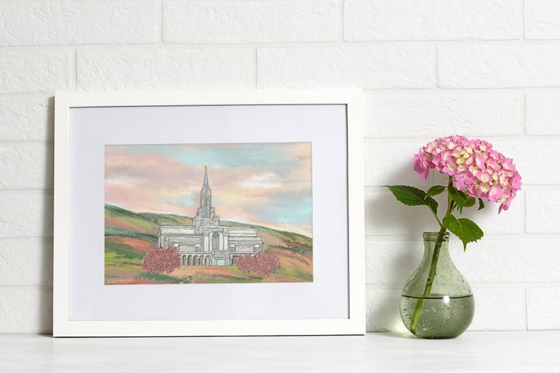 Peut inclure: Une impression encadr&eacute;e d'un temple blanc avec une fl&egrave;che, entour&eacute; d'arbres et de collines. Le ciel est rose et bleu p&acirc;le.